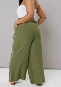 Olivgrön, vida byxor med elastisk midja och sidofickor. Har kontrasterande vit kant längs sömmarna. Mjuk tygtextur.