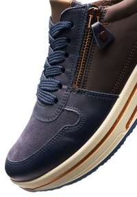 Marineblauwe sneaker met dikke veters, bruine enkelsluiting, ritssluiting aan de zijkant en een beige zool met een bronzen streepdetail.