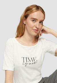 T-shirt blanc à manches courtes avec un col rond. Présente un texte imprimé "LE TEMPS est maintenant" en noir, avec un mélange de polices en gras et en italique.
