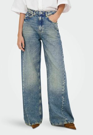 Brede blauwe denim jeans met een vervaagde was, vijf zakken en zichtbare stiksels. Het model draagt een losse witte blouse en bruine hakken.
