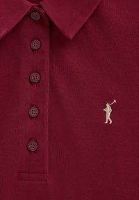 Polo bordeaux avec quatre boutons marron foncé et une figure blanche brodée tenant une maillette de polo sur la poitrine.