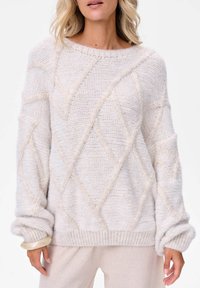 Kobieta ubrana w miękki, jasnobeżowy sweter w warkocze z diamentowym wzorem oraz długimi, lekko podwiniętymi rękawami, zestawiony z pasującymi spodniami.