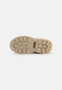 Lys beige fottøyssåle med et teksturert gummidesign, med dype spor og et heksagonalt mønster for godt grep. Merkeetikett synlig.