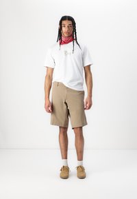 Pantaloni scurți bej cu o croială dreaptă; asortați cu un tricou alb cu text auriu; accesorizat cu o bandană roșie și adidași maron deschis.