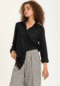 Black button-up shirt with a collar and long sleeves, paired with high-waisted, checkered patterned trousers. Smooth fabric texture.