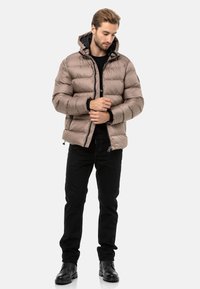 Hellbraune, gesteppte Jacke mit Kapuze, mit einem Reißverschluss vorne, gerippten Bündchen und schwarzem Futter. Kombiniert mit schwarzen Hosen und Stiefeln.