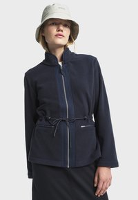 Chaqueta de forro polar azul marino con cuello alto, cierre de cremallera en el frente y cintura ajustable mediante cordón. Cuenta con dos bolsillos con cremallera y rayas verticales texturizadas.