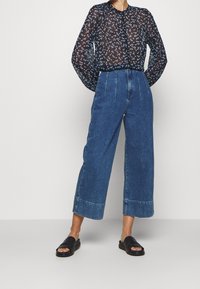 Schwarze durchsichtige Bluse mit blauem Blumenmuster, hochtaillierte, weit geschnittene blaue Jeans und schwarze Riemchensandalen.