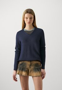 Navy V-ringad tröja i mjukt stickat tyg med långa ärmar, i kombination med en beige denim mini kjol som har ett urblekt mönster och ett fyrkantigt mönster.