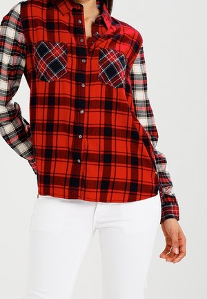 Czerwona koszula na guziki w kratę z czarno-białymi rękawami w kratę, dwiema kieszeniami na piersi, noszona z białymi spodniami na stojącej osobie.