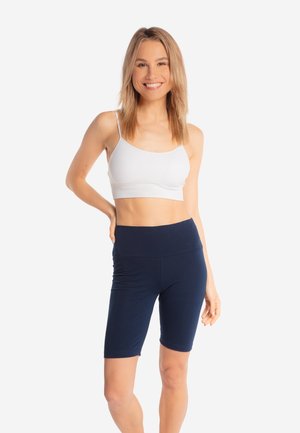 Młoda kobieta uśmiechająca się, ubrana w biały krótki top i granatowe, wysokowysokie krótkie spodenki rowerowe, stojąca z jedną ręką na biodrze.
