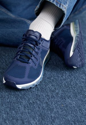 Granatowe buty sportowe z siateczkowym wzorem, noszone z białymi skarpetkami i niebieskimi dżinsami, pokazane na niebieskim wykładzinowym podłożu.
