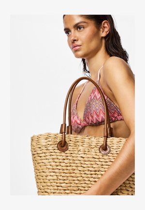 Mujer con top de bikini rosa con estampado que sostiene un gran bolso tote de paja tejido con asas de cuero marrón, mirando hacia la izquierda sobre un fondo liso.