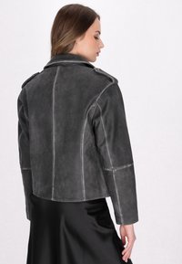 Grå læderjakke med kort snit, epauleter på skuldrene og kontrasterende hvid syning. Glat tekstur og tætsiddende design.