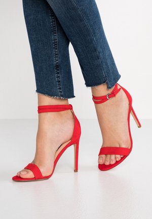 Røde ruskind højhælede sandaler med ankelstropper. Åben tå-design, slank hæl og glat tekstur. Båret med jeans med frynsede kanter.