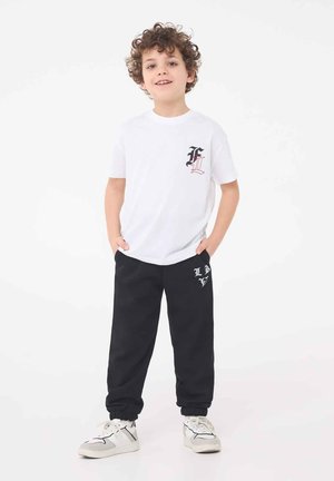 Maglietta in cotone bianca con logo rosso e nero sul petto, abbinata a pantaloni della tuta neri con grafica bianca, completata da sneakers chiare.