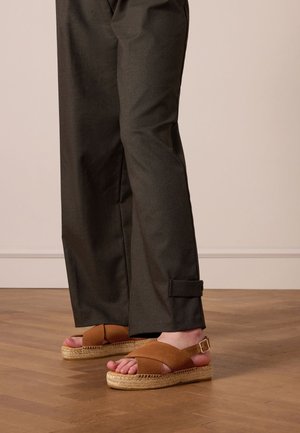 Brown espadrille sandals with cross-strap design and a woven jute sole, paired with dark, loose-fitting trousers featuring a side toggle.