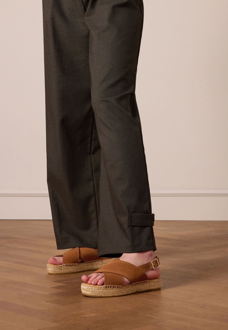 Sandales espadrilles marron avec un design à brides croisées et une semelle en jute tressé, associées à un pantalon sombre et ample avec un cordon sur le côté.