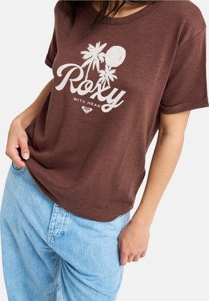 Persoon die een bruin Roxy T-shirt met een wit palmboomdesign en lichtblauwe spijkerbroek draagt, poseert met één hand in de zak.