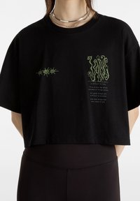 Černé zkrácené tričko vyrobené z bavlny, s grafickými designy v zelené barvě, včetně textu a vzorů hvězd na přední straně. Krátké rukávy.