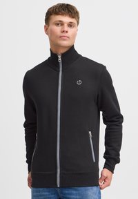 Schwarze Zip-Jacke mit hohem Kragen, aus weichem Material gefertigt. Verfügt über zwei seitliche Taschen mit Reißverschlüssen und einem Logo auf der Brust.