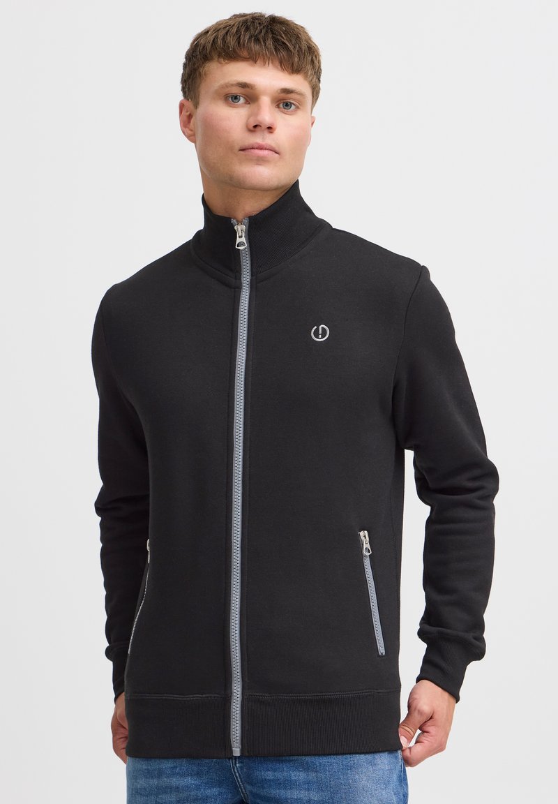 Schwarze Zip-Jacke mit hohem Kragen, aus weichem Material gefertigt. Verfügt über zwei seitliche Taschen mit Reißverschlüssen und einem Logo auf der Brust.