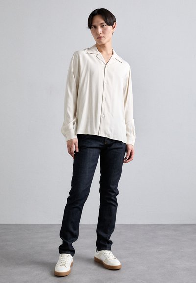 Licht beige overhemdd met een zachte textuur, gecombineerd met donkere spijkerbroek en witte sneakers met tan zolen. Staande pose voor een effen achtergrond.