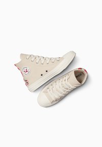 Zapatillas altas beige de lona, con puntera redonda, suela de goma blanca, ojales de metal y acentos florales rojos en el talón.