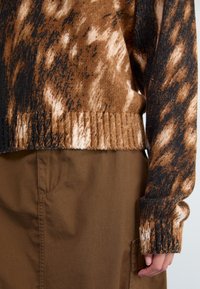 Knit sweater with brown, black, and cream color patterns. Ribbed cuffs and hem. Worn over a brown fabric with a side pocket detail.