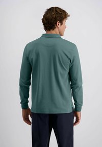 Mann mit lockigem Haar, der ein türkisfarbenes Langarmpolohemd und dunkle Hose trägt, von hinten stehend vor einem schlichten weißen Hintergrund.