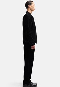 Czarna kurtka sztruksowa, dopasowane spodnie i błyszczące czarne buty. Wąski krój z zapięciem na guziki z przodu i bocznymi kieszeniami. Gładka faktura na całej powierzchni.