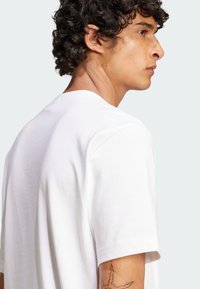 White cotton T-shirt with short sleeves, crew neck, and smooth texture. Features a subtle shoulder seam and a tattoo visible on the arm.