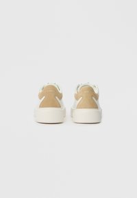 Ζευγάρι λευκών αθλητικών παπουτσιών με μπεζ σουέτ τμήματα στη φτέρνα, προβαλλόμενα από πίσω σε λείο ανοιχτό γκρίζο φόντο.