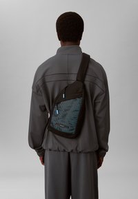 Bolso de bandolera negro con un panel de malla azul, que cuenta con un cierre con cremallera y una correa ajustable, llevado sobre un atuendo gris.