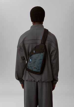 Zwarte schoudertas met een blauw gaaspaneel, voorzien van een ritsluiting en een verstelbare schouderband, gedragen over een grijze outfit.