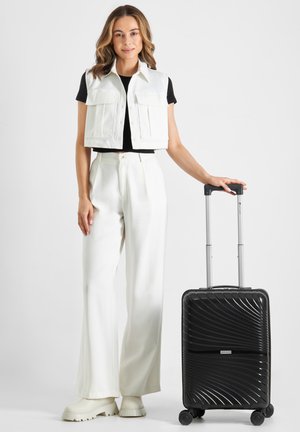 Black hard-shell suitcase with a wave pattern, silver handle, and four wheels. Model in white vest and pants, black top, and light boots.