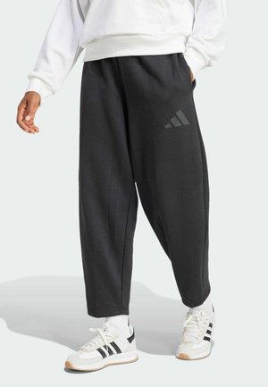 Personne portant un pantalon noir ample avec un petit logo sur la cuisse, associé à des baskets blanches avec des rayures noires et des chaussettes blanches.