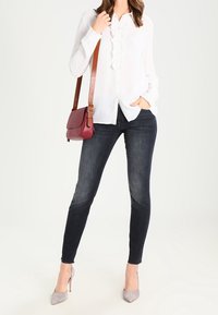 Camicia bianca con dettagli a volant, jeans skinny scuri, tacchi grigio chiaro e una borsa a tracolla bordeaux. Texture morbide e design aderente.