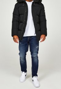 Svart vadderad jacka med en fuskpälsluva, dragkedjefickor, bärs över en vit skjorta, ihop med slitna blå jeans och vita sneakers.