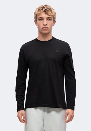 Giovane uomo con capelli biondi corti che indossa una semplice maglietta nera a maniche lunghe e pantaloni chiari, in piedi davanti a uno sfondo bianco.