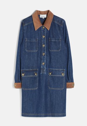 Kobieta w niebieskiej sukience z denimu z brązowym kołnierzem i mankietami, w połączeniu z wysokimi brązowymi skórzanymi butami, stoi na jednolitym tle.