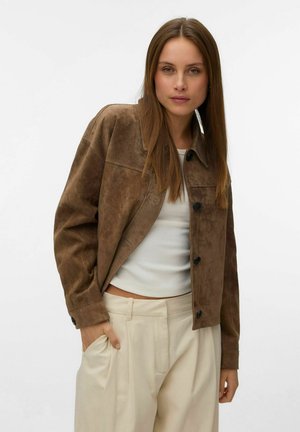 Jeune femme aux longs cheveux bruns portant une veste en daim marron, un top blanc et un pantalon plissé beige, une main dans la poche.