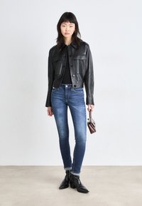 Μαύρο δερμάτινο κοντό μπουφάν με κουμπιά πατίνας, δύο τσέπες στο στήθος, συνδυασμένο με εφαρμοστά μπλε τζην και μαύρες μποτες αστραγάλου. Συμπεριλαμβάνεται καφέ τσάντα.