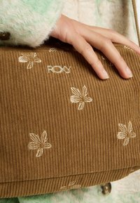 Braune Corduroy-Clutch mit floraler Stickerei und dem "ROXY"-Logo. Der Stoff hat eine strukturierte Ripp-Oberfläche. Eine Hand hält die Tasche.
