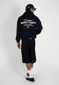 Homme portant un sweat à capuche bleu marine avec le texte « Prohibited Modern Yuppies », short bleu marine, chaussettes blanches, chaussures noires et casquette bleu marine, debout devant un fond blanc.
