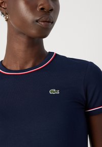 T-shirt ajusté bleu marine avec un col rond, des bordures rouges et blanches, et un logo de crocodile vert brodé sur la poitrine. Texture de tissu lisse.