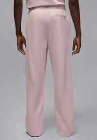 Henkilö, joka pukee vaaleanpunaiset kiristysnauhalla varustetut housut, joissa on takana vetoketjutasku, seisoo yksinkertaisen harmaan taustan edessä.