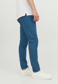 Slim-fit, blå stretchbyxor med synliga sömmar, avsmalnande ben och uppvikta fållar över vita sneakers. Materialet verkar mjukt och bomullsliknande.