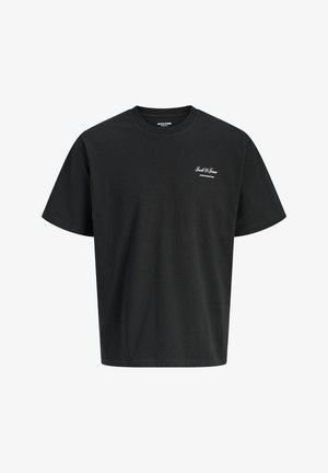 Χαμογελαστό αγόρι που στέκεται φορώντας μαύρο κοντομάνικο μπλουζάκι, ανοιχτό μπλε τζιν με φαρδιά μπατζάκια και λευκά αθλητικά παπούτσια, μπροστά σε απλό ανοιχτό φόντο.