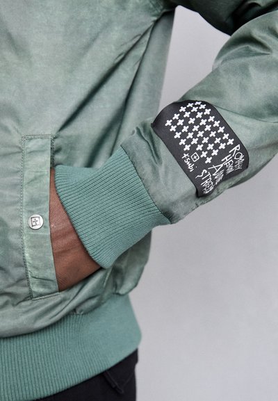 Veste bomber verte avec des poignets côtelés, présentant un patch noir avec un texte graphique blanc et des croix sur la manche. Détail de bouton à l'avant.